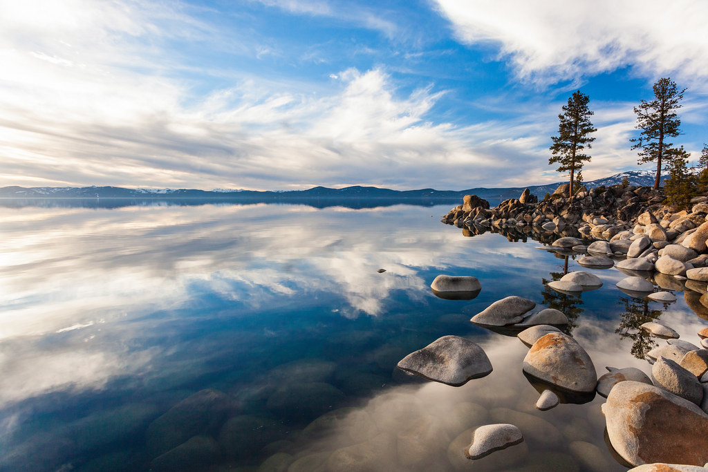 Paisagem serena de lago espelhado com rochas e pinheiros, refletindo a calma interior necessária para acalmar o crítico 